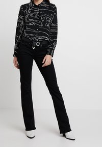 Camisa negra de botones con un patrón de arte lineal blanco, combinada con pantalones acampanados negros y un cinturón con una hebilla metálica circular.
