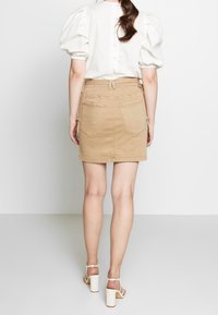Jupe en denim beige au design ajusté, avec poches arrière, taille ajustable et texture lisse, associée à un haut à manches bouffantes blanches.