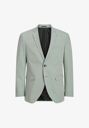 Dreng iført en lys grøn dragt med sorte knapper, hvid skjorte med krave og beige sneakers, stående mod en lys baggrund.