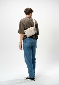Borsa a tracolla beige con più scomparti con zip, materiale morbido, tracolla regolabile, indossata su una camicia marrone ampia e jeans blu.