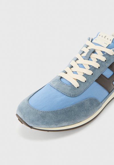 Baskets en daim bleu clair et tissu avec des lacets crème et un logo latéral marron foncé sur une semelle blanche, sur fond gris.