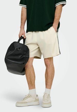 Man draagt beige shorts, witte sneakers en een zwart T-shirt en houdt een zwarte leren rugzak vast bij het handvat.