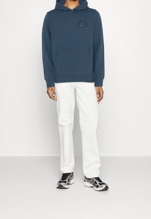 Mörkblå bomulls-huvtröja med huva och framficka samt svart logotyp; matchad med vita manchesterbyxor och svarta sneakers.