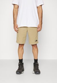 Man som bär beige knälånga shorts med The North Face-logotyp, svarta strumpor, svarta vandringsskor och en vanlig vit T-shirt.