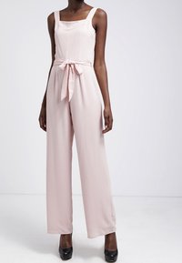 Vaaleanpunainen haalarimaha, jossa on neliökaula-aukko, leveät hihnat ja solmittava vyötärö. Kankaan pinta on sileä; löysät housut ulottuvat lattiaan. Mustat korot on käytössä.