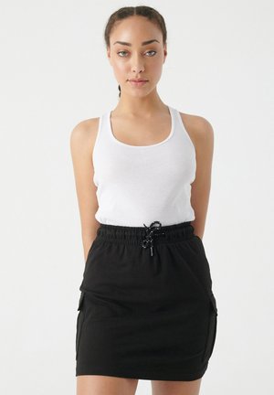 Joven mujer con trenzas lleva una camiseta sin mangas blanca y una falda negra con cordón, de pie frente a un fondo claro y liso.