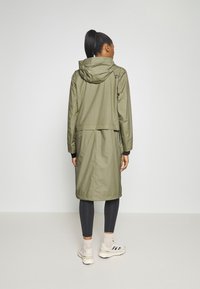 Oliwkowa, wodoodporna kurtka z kapturem, warstwowe wzornictwo i elastyczne mankiety; w zestawieniu z ciemnymi legginsami i jasnymi butami sportowymi.