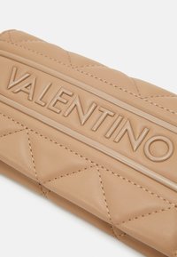 Bežová kožená peňaženka s prešívaným vzorom a vytlačeným logom "VALENTINO" vpredu. Obsahuje prešívané detaily a hladký, textúrovaný povrch.