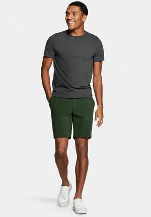 Man glimlacht, loopt vooruit en draagt een donkergrijs T-shirt met korte mouwen, donkergroene korte broek en witte sneakers, handen gedeeltelijk in de zakken.