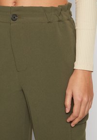 Detailní záběr pasu a ruky oblečené v olivově zelených kalhotách s vysokým pasem s knoflíkem a viditelném béžovém tričku s dlouhým rukávem.