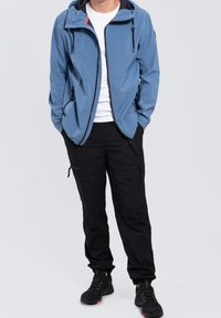 Chaqueta ligera azul claro con capucha, acentos negros y bolsillos con cremallera, combinada con pantalones cargo negros y calzado negro.