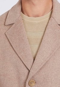 Beige uldfrakke med en enkelt knap over en cremefarvet striktrøje med vandrette strukturerede striber.