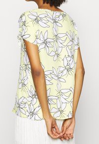 Kortærmet top i lys gul stof med blomsterprint i sort og hvid. Inkluderer samling ved ærmeender.