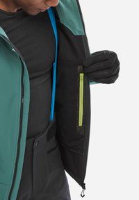 Person wearing black gloves opens teal jacket revealing black mesh inner pocket with a vertical yellow zipper.