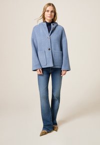 Chaqueta de mezcla de lana texturizada de color azul claro con capucha, dos bolsillos frontales y grandes botones. Combinada con jeans ajustados y zapatos de diseño.