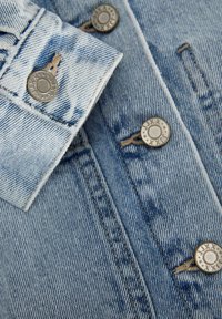 Tessuto in denim azzurro chiaro con una superficie testurizzata. Presenta bottoni e dettagli di cucitura in tono argento. Il colletto è piegato, mostrando le linee di cucitura.