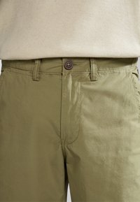 Pantalones verde oliva hechos de una tela ligera con una textura suave. Presentan un cierre de botón y detalles cosidos en la cintura y los bolsillos.