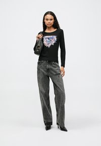 Moteris vilki juodą ilgą rankovę marškinėlį su spalvinga grafika, pilkas plačių kojų džinsus, juodas aukštakulnius batus ir laiko juodą rankinę ant peties.