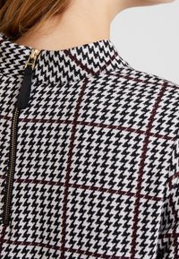 Gros plan sur un vêtement à motif pied-de-poule avec des lignes de grille rouges et une fermeture éclair dorée au niveau de l'encolure arrière, porté par une personne à la peau claire.