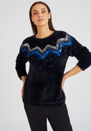 Donkerblauwe fluffy trui met een zilveren en blauwe pailletten zigzagpatroon over de borst. Lange mouwen en ronde halslijn. Zachte textuur.
