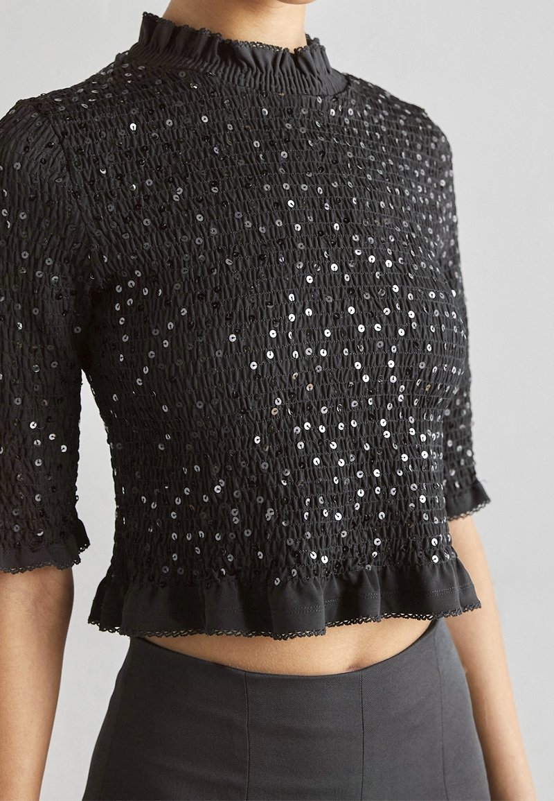 Top court noir avec texture froncée et sequins brillants dispersés, col haut volanté et manches courtes sur une personne.