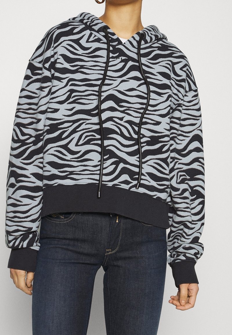 Femme portant un sweat à capuche rayé zèbre noir et gris avec des cordons noirs et un jean en denim foncé, debout devant un fond uni.