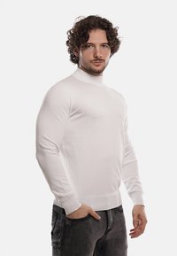 Maglione bianco a collo alto a maglia con maniche lunghe e polsini a coste, abbinato a jeans scuri, mostrato su un modello contro uno sfondo semplice.