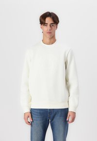 Sudadera blanca con cuello redondo, logo en relieve en el pecho y mangas largas, combinada con pantalones vaqueros azul denim. Textura de tela suave.