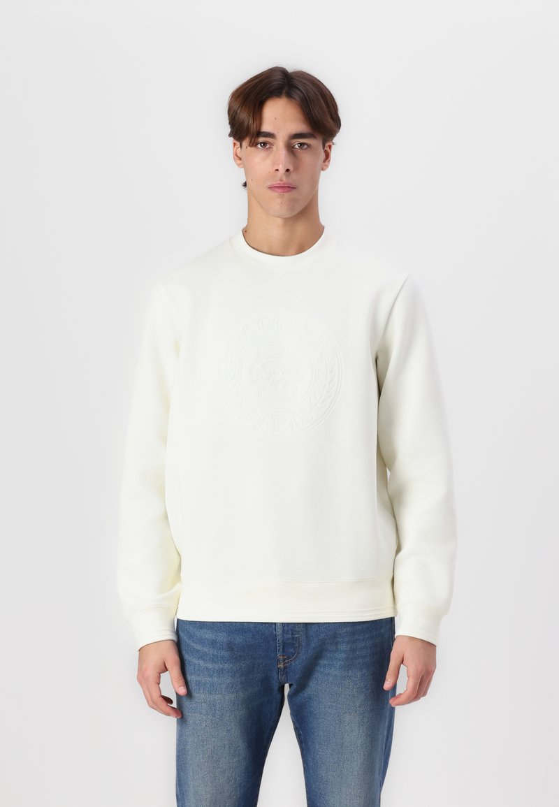 Sudadera blanca con cuello redondo, logo en relieve en el pecho y mangas largas, combinada con pantalones vaqueros azul denim. Textura de tela suave.