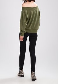 Olivgrön off-shoulder sweatshirt, oversize passform, kombinerad med svarta skinnyjeans och leopardmönstrade ankelstövlar, sedd bakifrån.