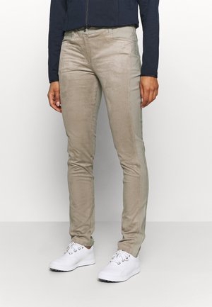 Persoon draagt beige slim-fit broek, witte sneakers en een donkerblauw jack met rits, staand tegen een effen lichte achtergrond.