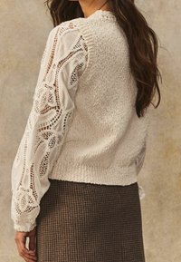 Biała dzianinowa bluzka z koronkowymi detalami na bufiastych rękawach, z teksturowanej tkaniny oraz okrągłym dekoltem, w zestawie z brązową spódnicą w kratkę.