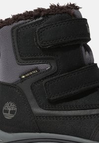 Svart och grå vattentät stövel med kardborreband, fleecekant och en GORE-TEX-etikett. Har en logotyp på sidan och texturerade tyginslag.