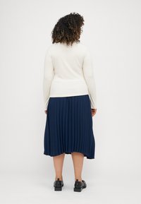 Ljust beige ribbat långärmad topp i kombination med en mörkblå plisserad midkjol, med svarta ankelstövlar, sedd bakifrån.