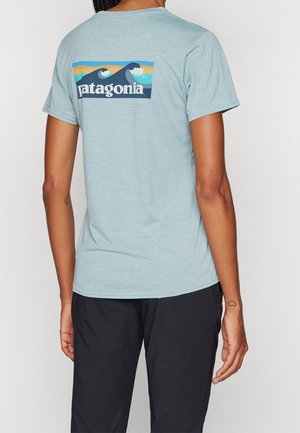 Persoon die een lichtblauw Patagonia T-shirt met golflogo op de rug en een zwarte broek draagt, staand tegen een effen achtergrond.