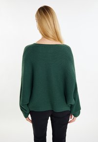 Zöld csíkos kötött pulóver, oversize fazonban, széles, hajókivágású nyakkal, ejtett vállakkal és hosszú ujjakkal, fekete nadrággal párosítva.