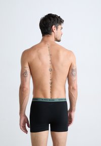 Homme avec un tatouage de colonne vertébrale géométrique et des tatouages sur les bras portant des slips Björn Borg noirs, se tenant devant un fond blanc uni.