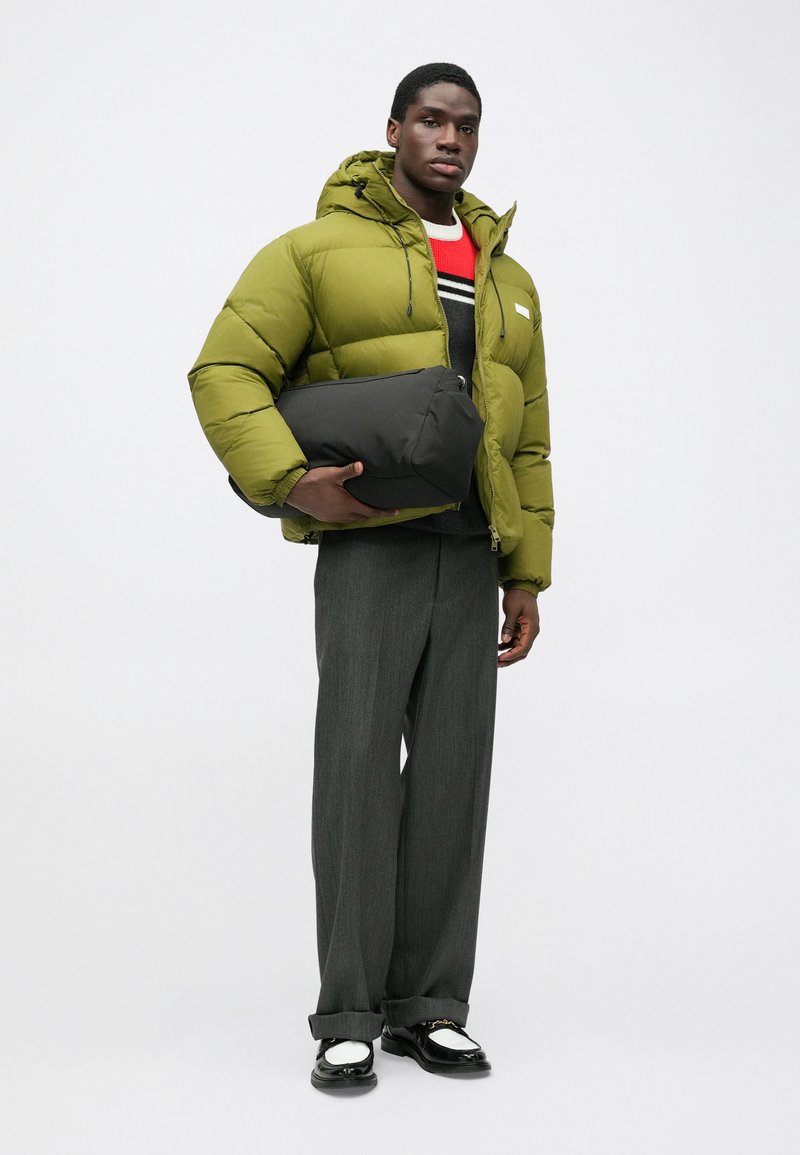 Green puffer jacket with a hood, holding a black bag. Grey, textured, wide-legged trousers and black and white shoes with accents.