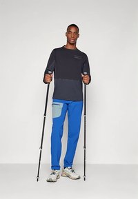 Muškarac u crno-plavoj aktivnoj odjeći drži dva planinarska štapa pred jednostavnom bijelom pozadinom.