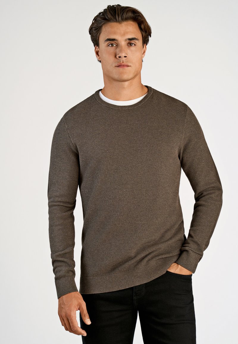 Ruskea neulepusero, jossa on pyöreä kaula-aukko ja pitkät hihat, sisältää tekstuuripinnan ja ribbireunat, yhdistettynä mustiin farkkuihin.