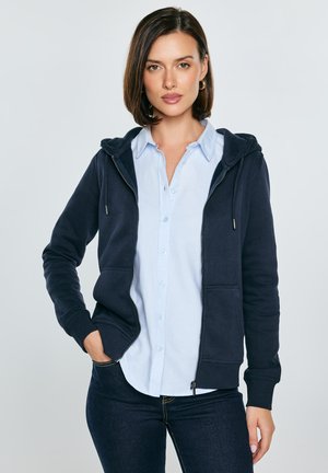 Frau mit braunen Haaren, die ein hellblaues Hemd mit Knöpfen, einen marineblauen Kapuzenpullover mit Reißverschluss und dunkle Jeans trägt, steht mit einer Hand in der Tasche.