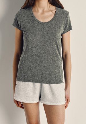 Kvinde iført en mørkegrå t-shirt med korte ærmer og lysegrå shorts, stående foran en ensfarvet neutral baggrund.