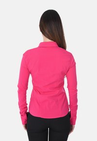 Camicia aderente a maniche lunghe di colore rosa acceso con colletto. Tessuto texturizzato, cuciture dettagliate e polsini con bottoni. Indossata con pantaloni neri.