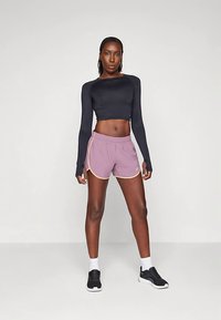 Sportiška moteris stovi su rankomis šonuose, vilkėdama juodą ilgesnį rankovėmis marškinėlį, mauvinius bėgimo šortus, baltas kojines ir juodus sportinius batelius ant balto fono.