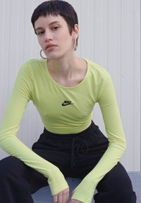 Hosszú ujjú, világos zöld Nike felső, testhezálló fazonnal. Kerek nyakkivágással és fekete logóval rendelkezik. Fekete nadrággal párosítva. Sima textúrájú.
