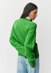 Zaļš adīts džemperis ar ribotu apakšmalu un piedurknēm, ar apaļu kakla izgriezumu. Gluda tekstūra, brīvs piegriezums. Uzvilkts virs augstām jostasvietām džinsiem.