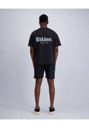 Mann steht mit dem Rücken zur Kamera, trägt ein schwarzes T-Shirt mit weißem Text „Blckbnns Design Dept.“ und schwarze Shorts, vor einfarbig hellem grauem Hintergrund.
