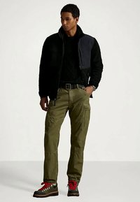 Άνδρας ντυμένος με μαύρη φλις ζακέτα, μαύρο πουλόβερ, ελιά χρώματος cargo παντελόνι και μαύρες μπότες με κόκκινα κορδόνια, στέκεται με το ένα χέρι στην τσέπη.