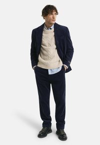 Marineblauer Cord Anzug mit einem strukturierten, cremefarbenen Kabelstrickpullover darunter. Kragenhemd am unteren Ende sichtbar, kombiniert mit schwarzen Schnürstiefeln.