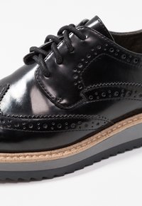 Chaussure oxford en cuir noir avec des perforations décoratives, bout rond et semelle en caoutchouc texturée avec un léger talon surélevé.